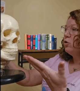 Doreen is talking to a skull that she is holding in her right hand. No human is attached to the skull currently, and it looks like it's plastic. She is reciting "to Polish or not to Polish, that is the question" to the skull, but since this is a picture, you can't hear her. She is wearing a pink tshirt and glasses and she is in a yellow room. There is a bookshelf behind her that used to have sanitizing wipes that she forgot to remove before taking the picture, so we put fake books on the shelf instead to make it look like we did it on purpose.
